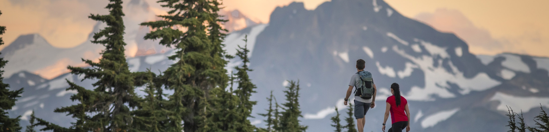 A man and woman stand on a rock looking towards mountains as the sun sets