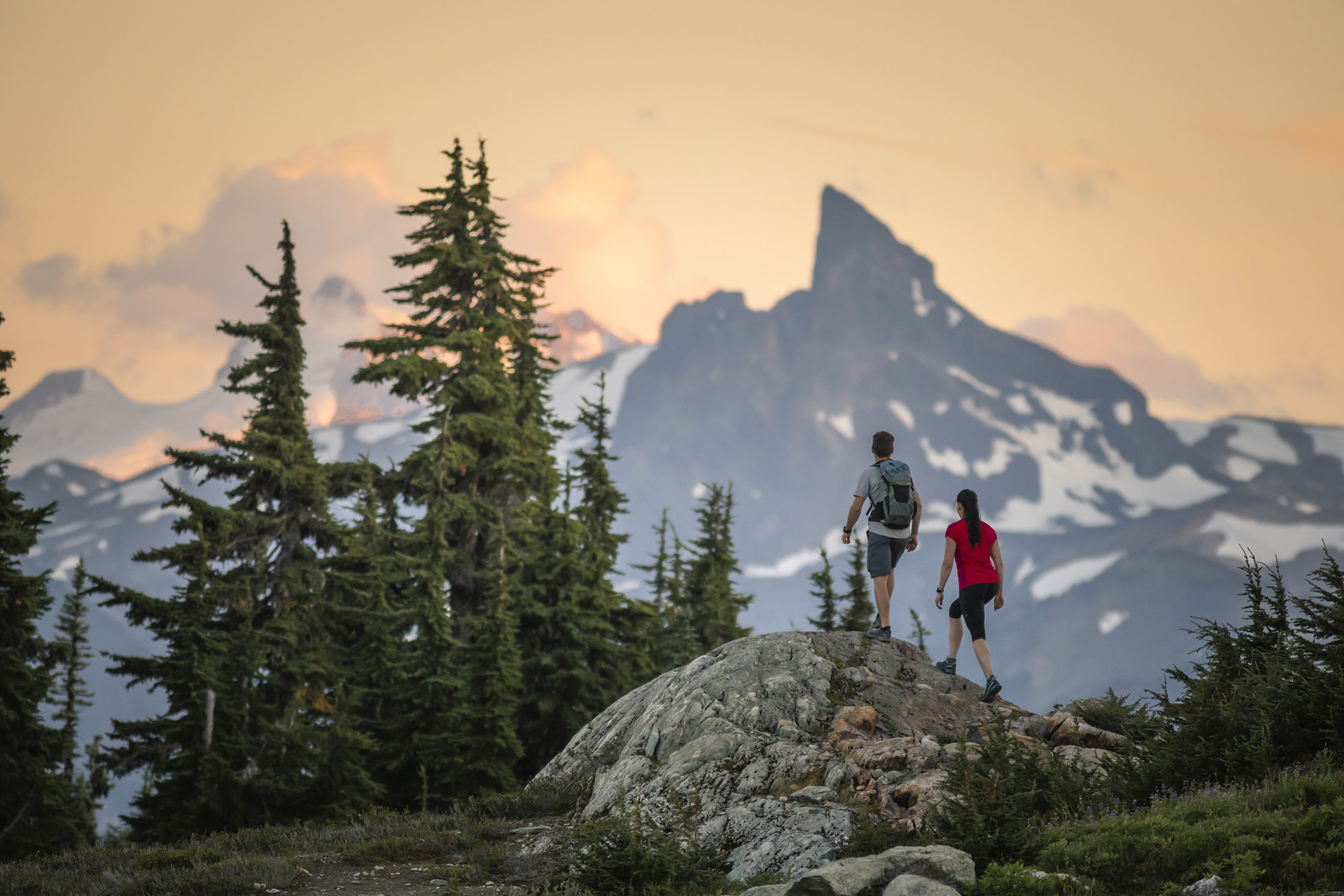 A man and woman stand on a rock looking towards mountains as the sun sets