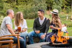 A group of friends sit around a fire during the day wih drinks