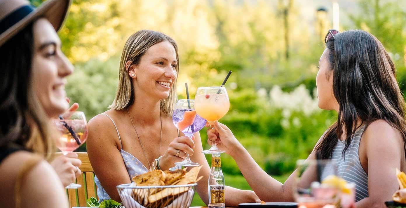 Two women cheers their drinks at an outdoor table of food
