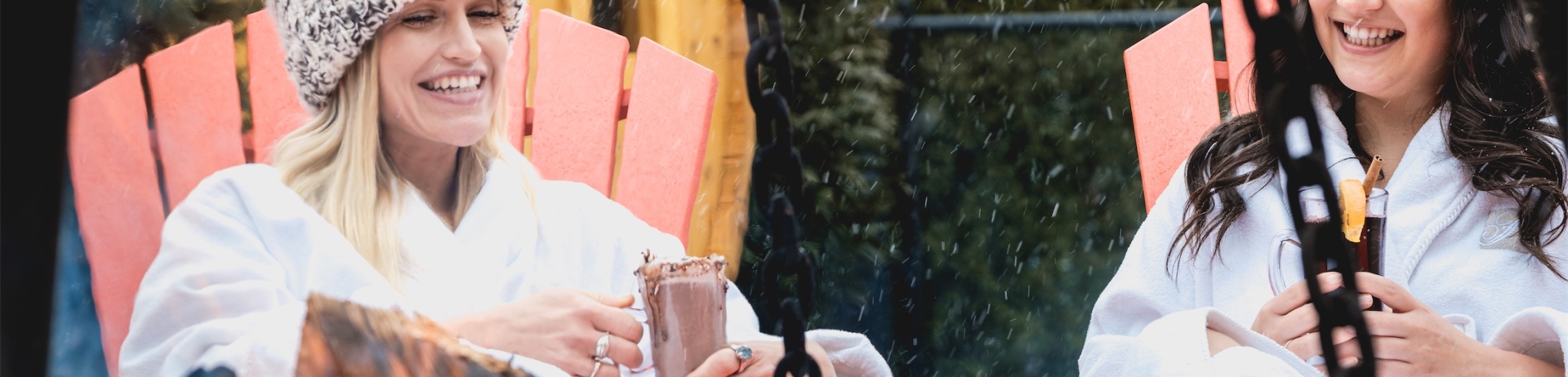two women in robes sitting in red chairs drinking cocktails next to an outdoor fire
