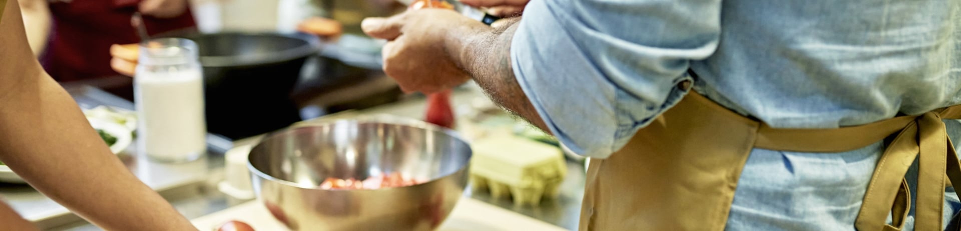 Close-up of men in aprons standing at cutting board slicing plum tomatoes as female colleagues and senior chef instructor work in background.
