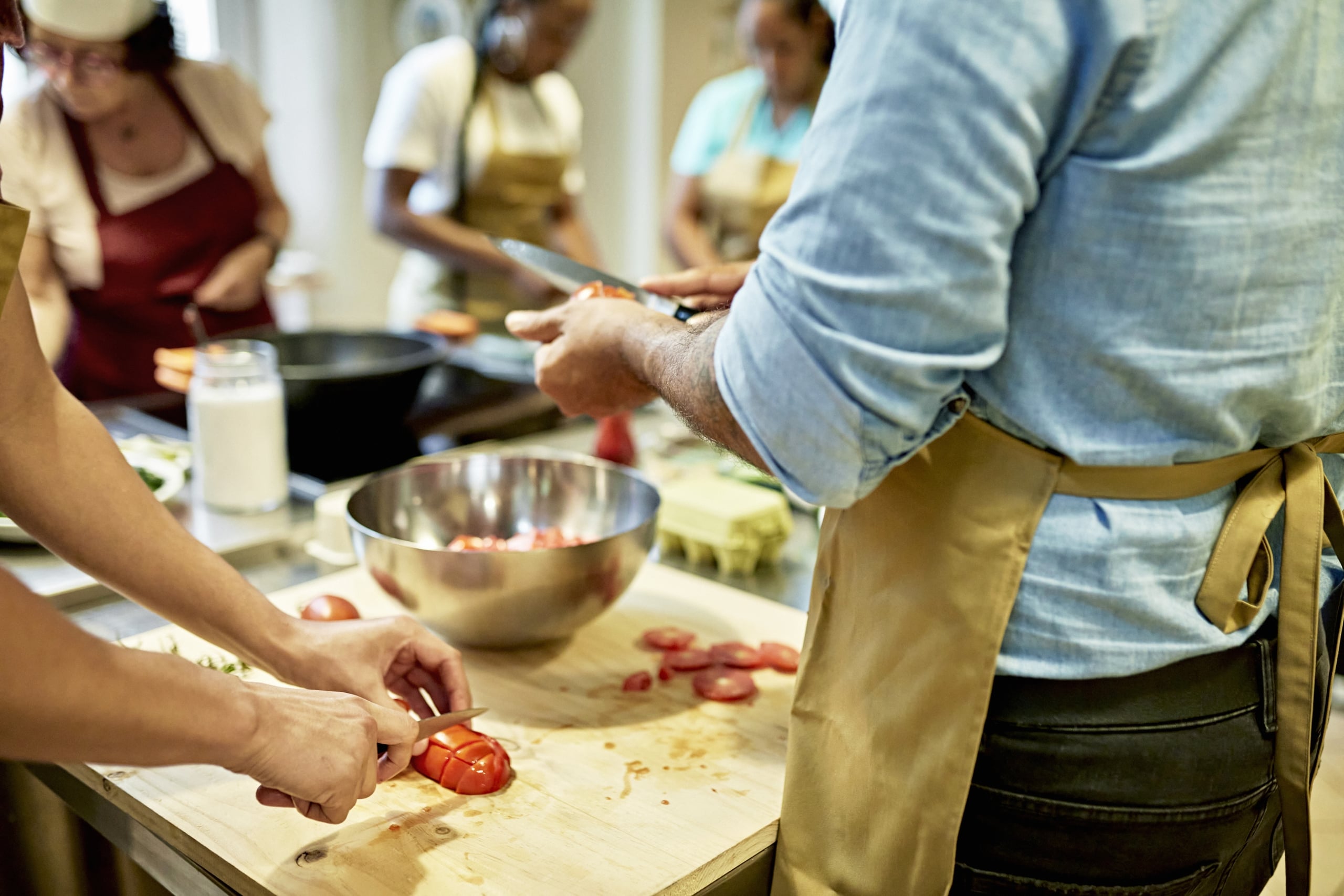 Close-up of men in aprons standing at cutting board slicing plum tomatoes as female colleagues and senior chef instructor work in background.