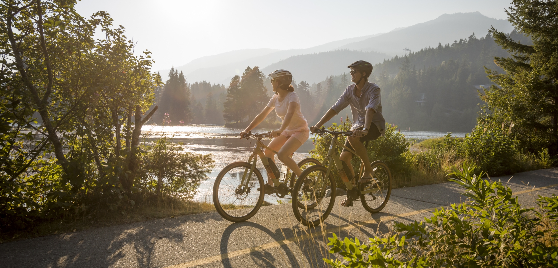 two people biking on a paved trail next to a lake in a mountain landscape