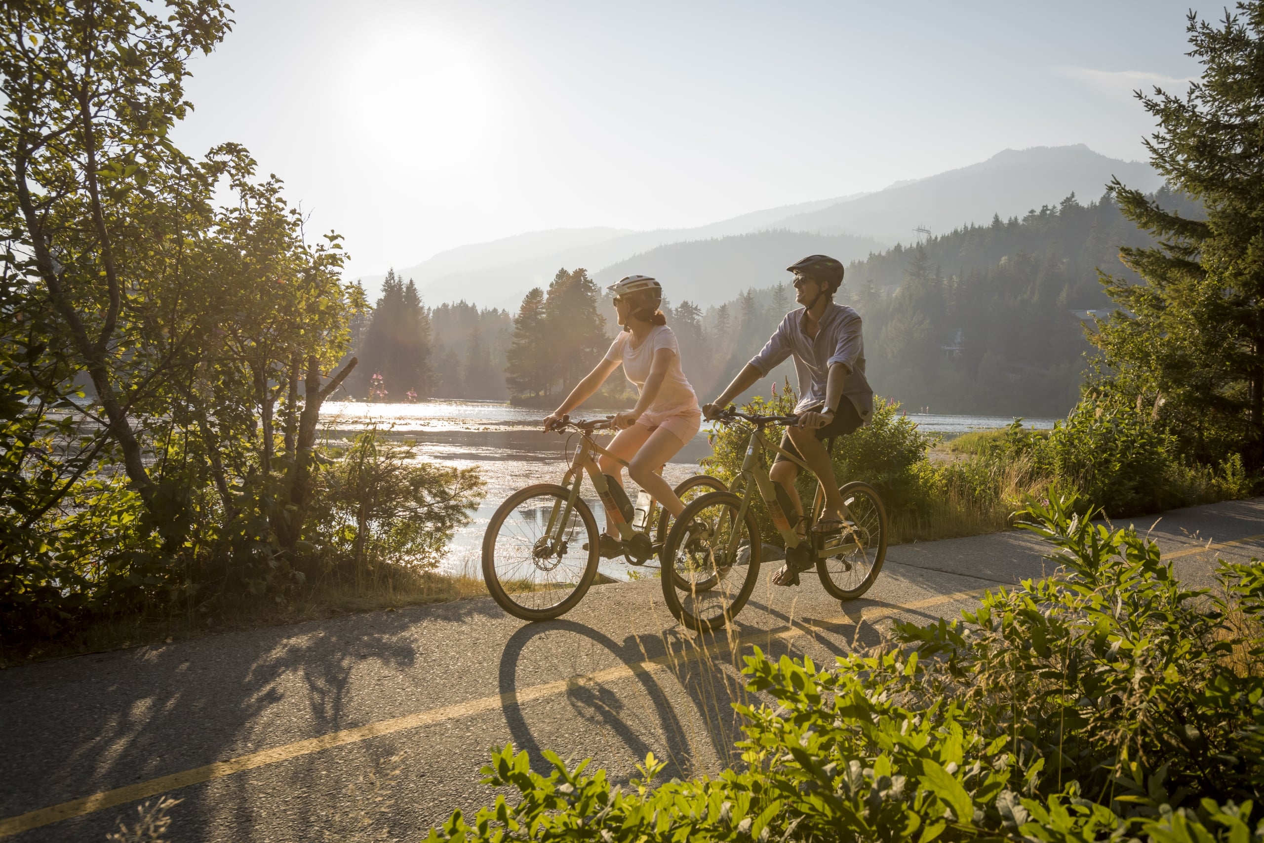 two people biking on a paved trail next to a lake in a mountain landscape